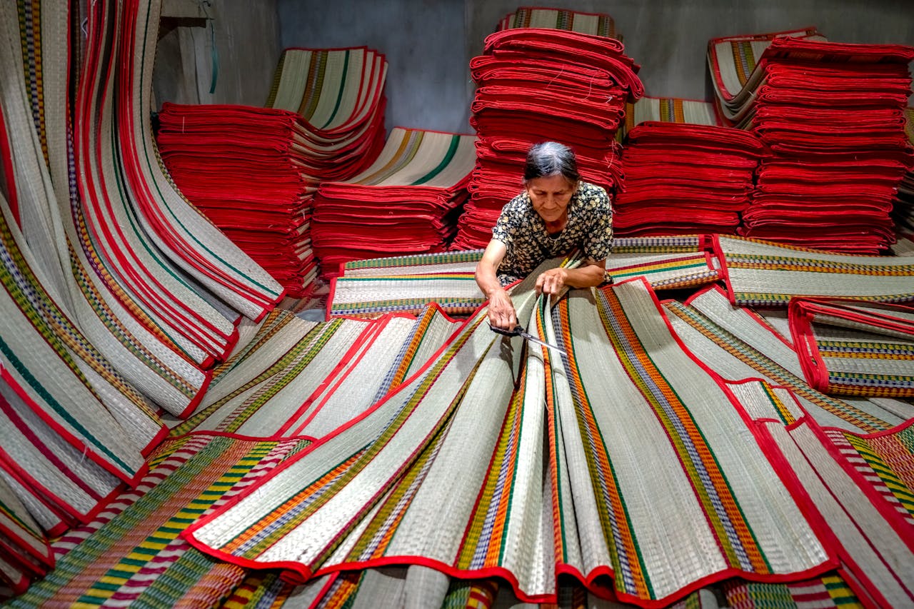 An elderly woman skillfully weaving colorful mats in a busy workshop setting.